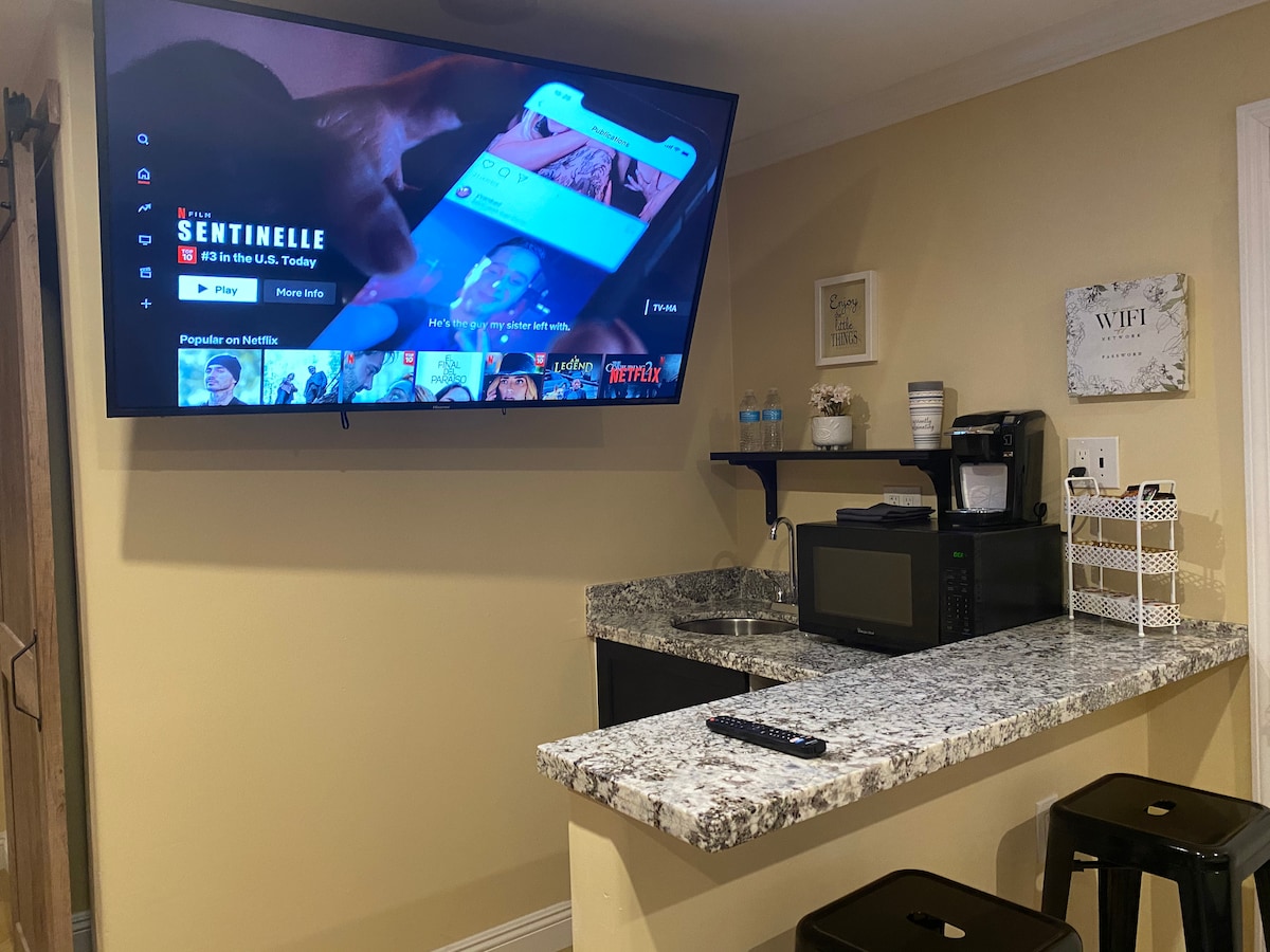 A flat-screen TV is mounted on the wall, displaying a streaming service menu. A kitchen area with a granite countertop holds a microwave and a coffee maker, alongside several bottles of water. Two bar stools are set at the counter for seating.