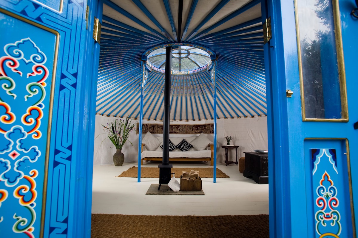 The entrance to a yurt reveals a spacious interior featuring intricately painted blue doors. A central wood-burning stove is surrounded by a natural fiber mat, with cozy seating and a decorative table. Natural light filters through a circular window at the top, enhancing the inviting atmosphere.