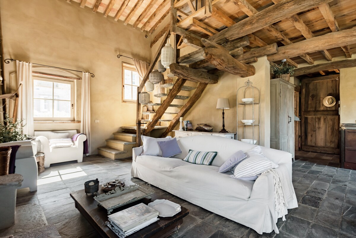 A cozy living room is highlighted by a spacious white sofa adorned with various patterned cushions. An elegantly designed wooden staircase ascends to the upper floor, while warm light filters through a window, illuminating the rustic stone floor and antique furniture.
