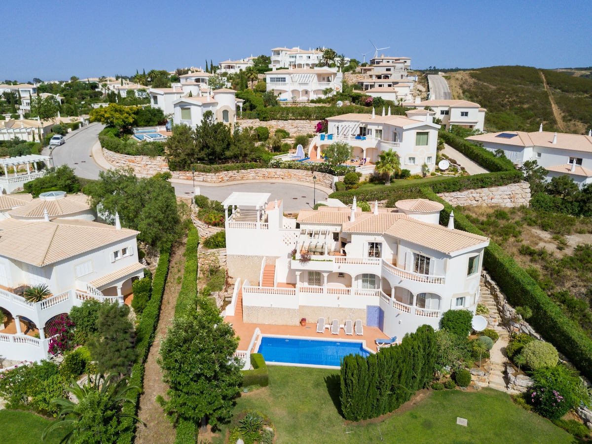 This aerial view showcases a spacious villa within a beautifully landscaped area. The large blue pool is surrounded by green hedges and lounge chairs, while the surrounding properties are visible on the hillside, contributing to the tranquil setting.