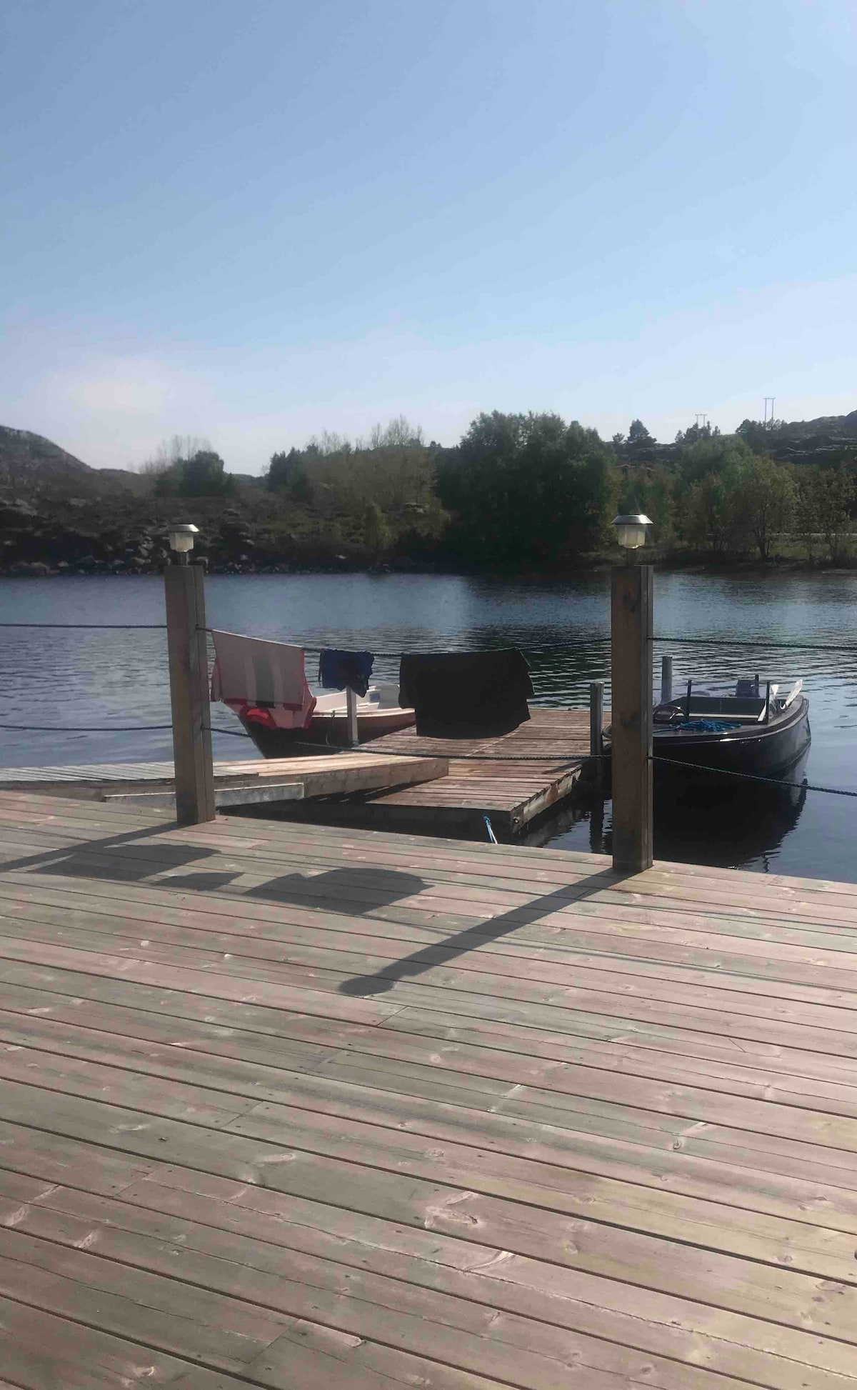 A wooden dock extends over calm water, featuring two chairs positioned for relaxation. A small boat is moored alongside, with green trees visible in the background. Sunlight reflects off the water's surface, creating a serene atmosphere.