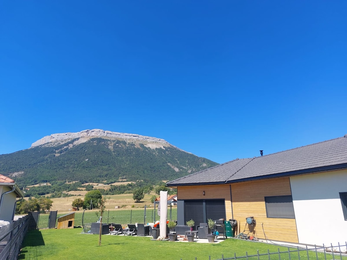 The image captures a clear blue sky above a modern house, surrounded by a well-maintained lawn. In the background, the mountainous landscape of Céüse is visible, showcasing its rugged terrain and greenery.
