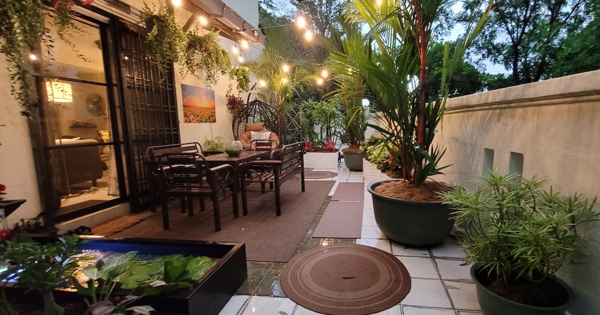 An outdoor patio area is featured with a dining table surrounded by seating, adorned with hanging plants and string lights. A lotus pond is visible alongside potted greenery, creating a relaxed environment for outdoor gatherings.