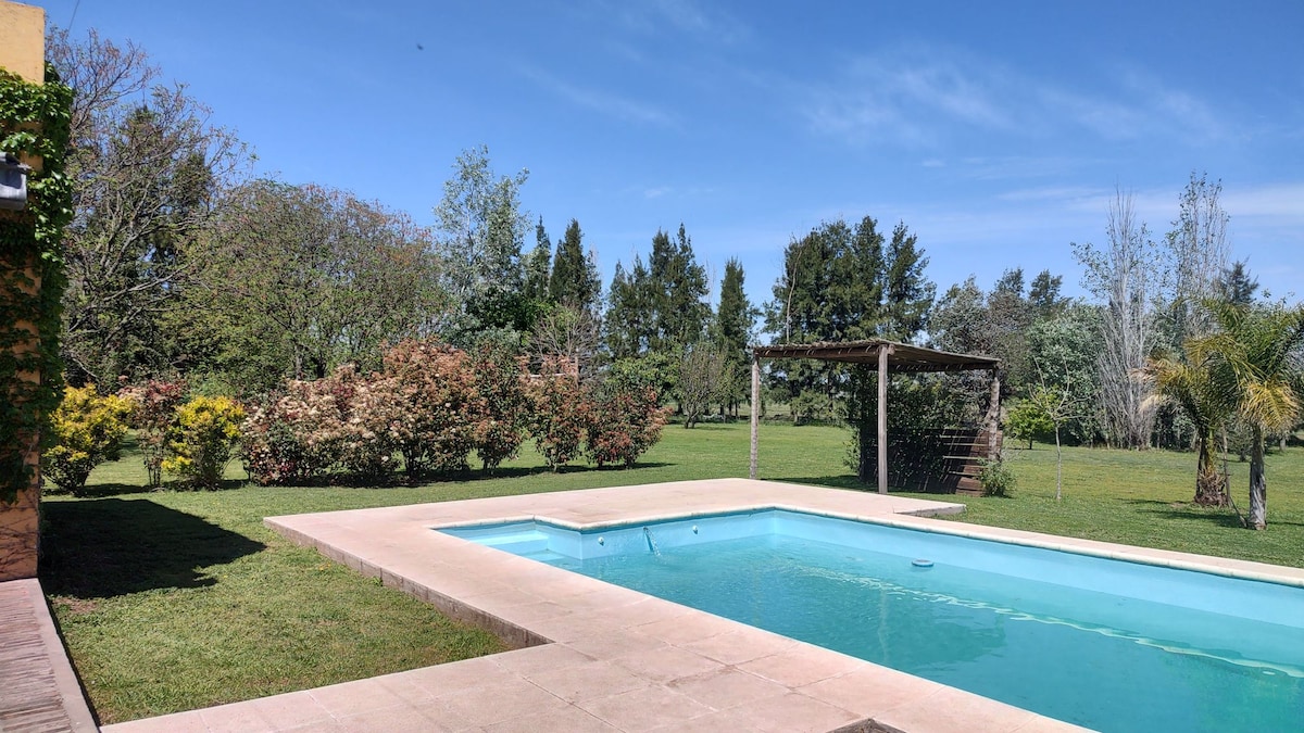 A serene outdoor area is featured, showcasing a clear swimming pool surrounded by lush greenery and blooming shrubs. A wooden pergola provides shaded seating, while trees frame the expansive lawn, offering a peaceful retreat in a natural setting.