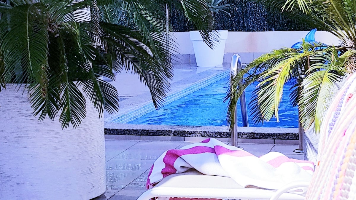 A close-up view of a private pool area is shown, framed by lush greenery. A lounge chair is positioned nearby, topped with a striped towel. The blue water in the pool sparkles under sunlight, creating a serene atmosphere.