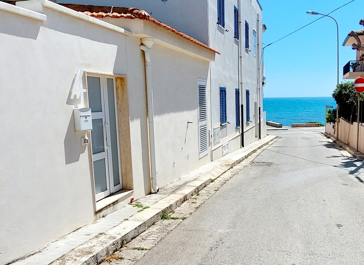 A view of a quiet street leads to the coastline, with a clear glimpse of the ocean in the background. The single-story building features light-colored walls and blue window shutters, while the well-maintained sidewalk offers an unobstructed path towards the water.