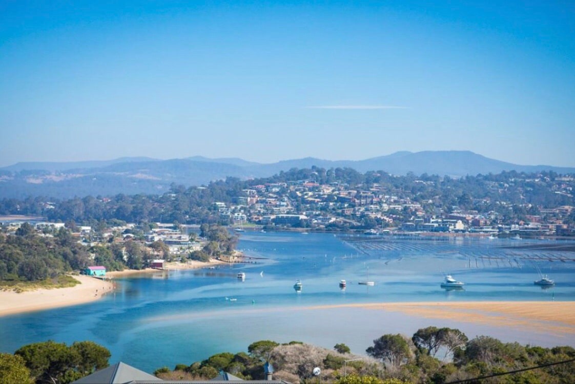 A sweeping view of a coastal town is captured, featuring calm waters reflecting the sky. Boats are seen moored in the bay, with hills in the distance creating a scenic backdrop. The landscape includes patches of greenery along the shore, adding a natural element to the vista.