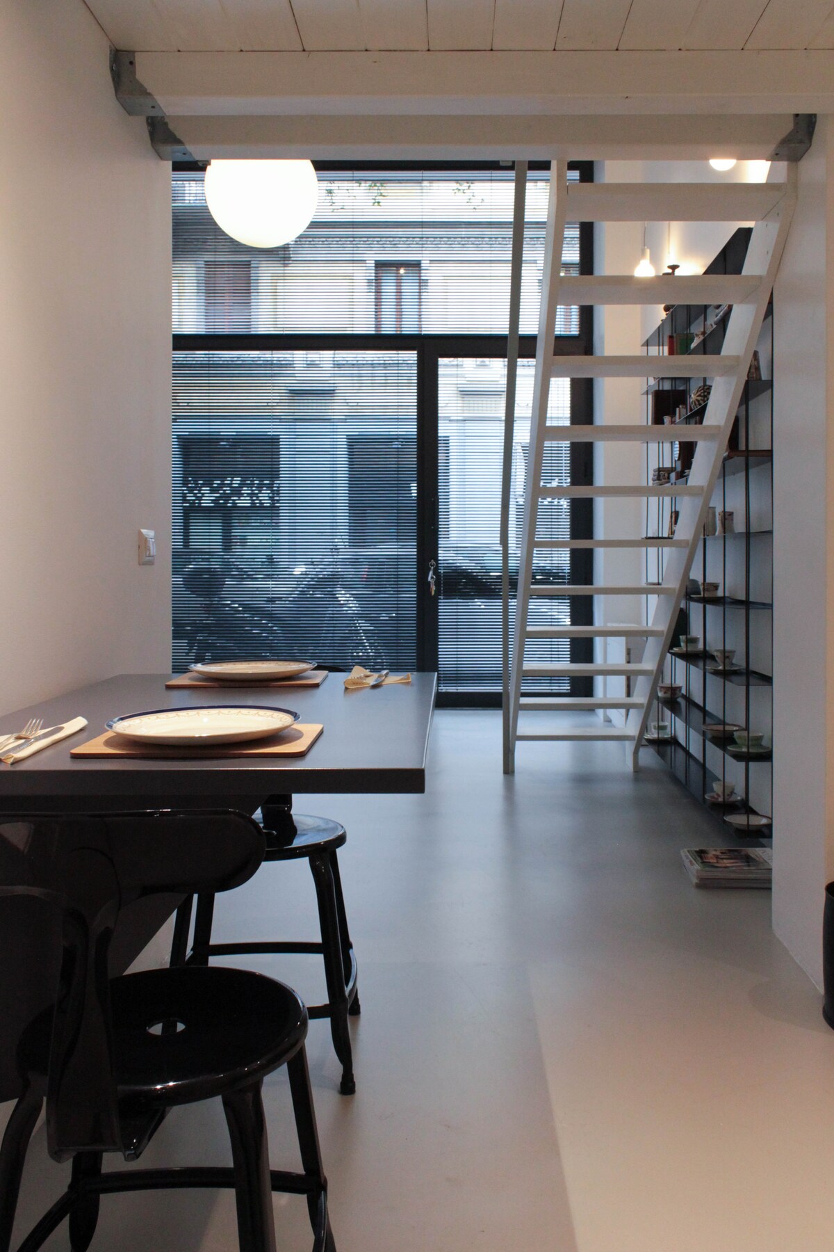 A sleek dining area features a minimalist table set for two, accompanied by black stools. A modern, open staircase leads to the upper level. Large glass doors provide an unobstructed view of the street outside, allowing natural light to fill the space.