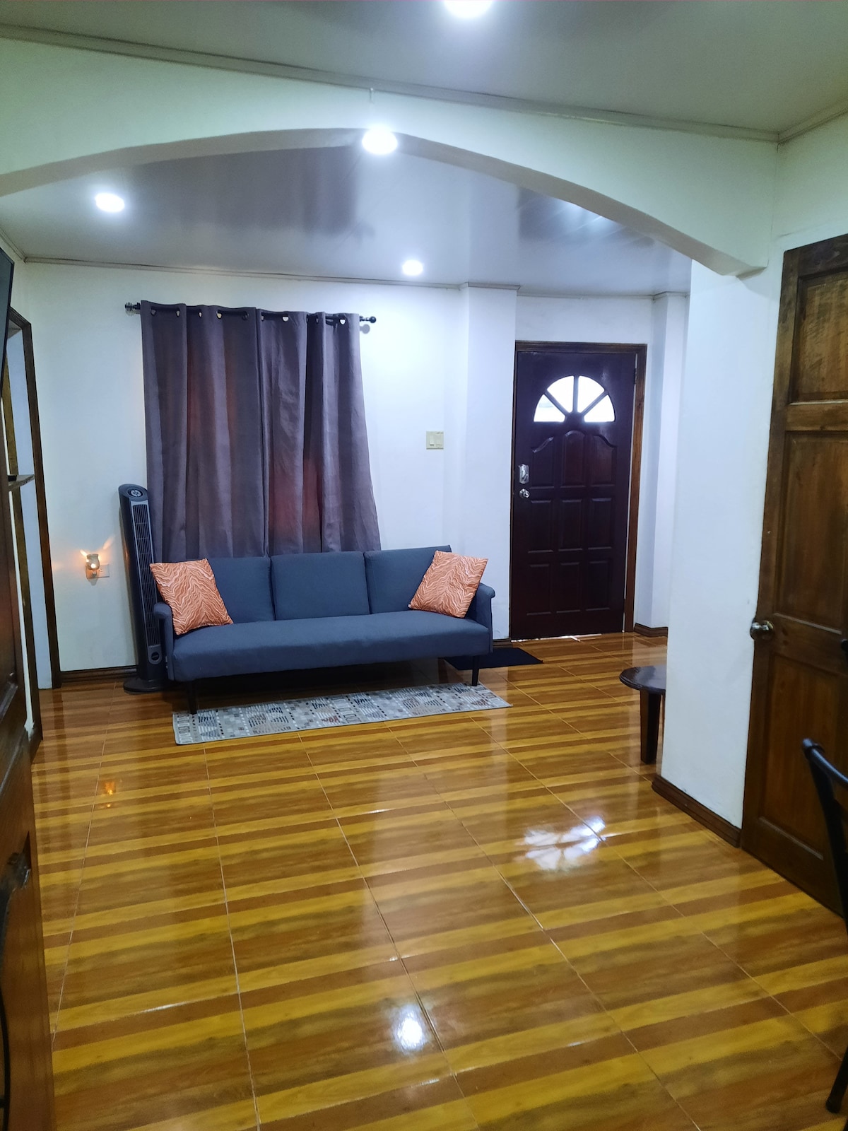 A cozy living area features a modern sofa adorned with orange accent pillows. Polished wooden flooring reflects warm light from recessed ceiling fixtures. A decorative rug anchors the space, while a dark curtain frames the window next to the entrance door.