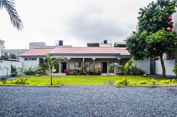 A charming guest house features a red-roofed design, surrounded by a neatly landscaped garden with lush greenery. Gravel pathways lead to the separate entrances of the two furnished apartments, offering a welcoming and serene outdoor space.