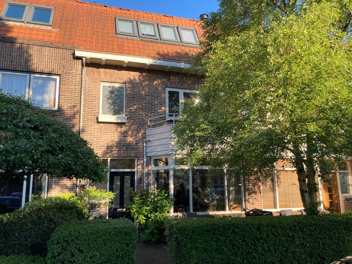 A spacious house is visible with a brick facade and a bright red tiled roof. Large windows allow natural light into the living areas. A well-kept garden with greenery frames the front, contributing to a welcoming exterior. A small terrace can be seen at the upper level.