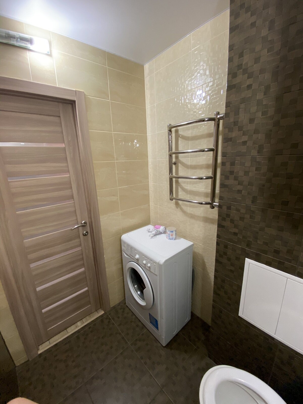 A compact bathroom is showcased, featuring a washing machine against a light-colored tiled wall. A modern towel rack is mounted alongside the wall, and a neutral door leads to the room. A toilet is positioned in the corner, maintaining a tidy and functional design.