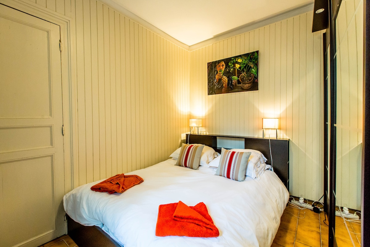 A cozy bedroom features a spacious bed with colorful accent pillows and neatly folded towels. Two bedside lamps provide soft lighting, and a piece of wall art adds a touch of decor. A mirrored wardrobe reflects the room's inviting atmosphere.