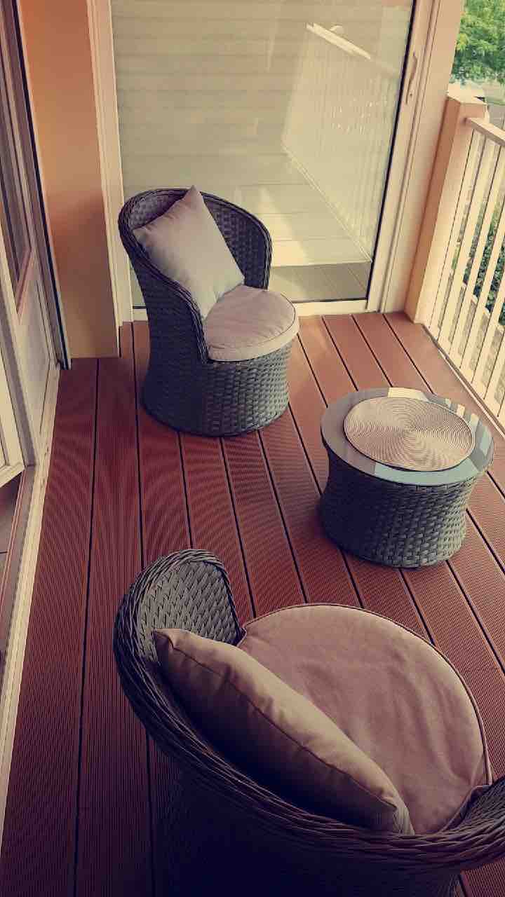A small balcony features two woven chairs with plush cushions and a round side table. Natural light illuminates the space, enhancing the wooden flooring and providing a cozy spot for relaxation.
