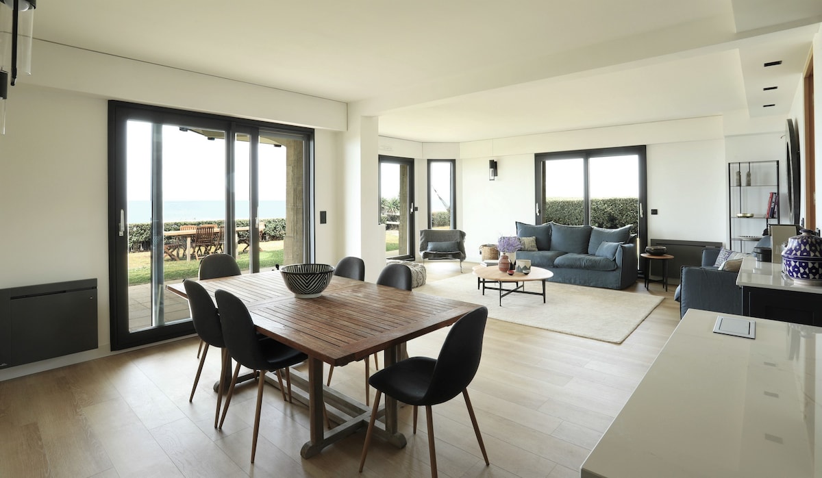 A spacious living area features a wooden dining table surrounded by black chairs, with large glass sliding doors providing panoramic views of the sea. A comfortable seating arrangement is positioned nearby, complemented by a neutral rug and natural light illuminating the space.