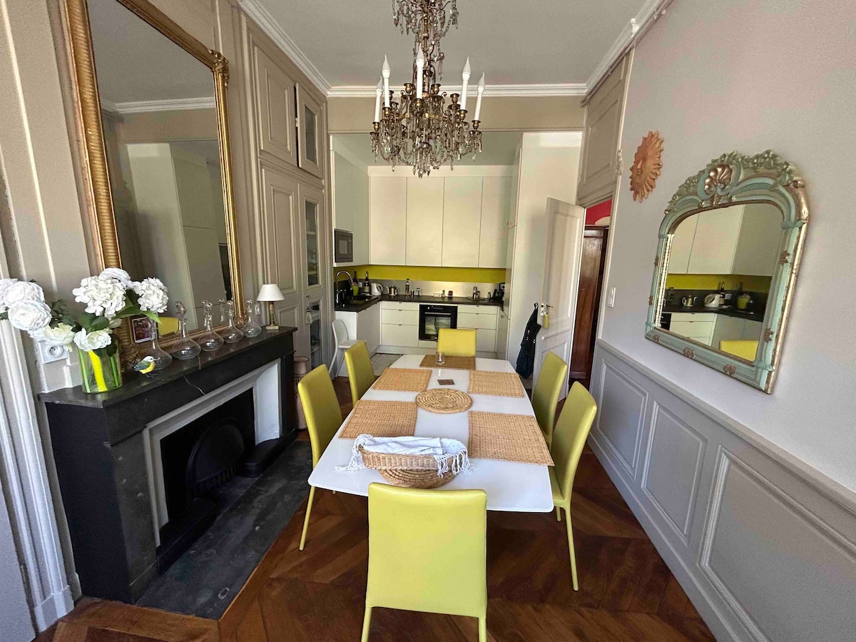 A spacious dining area features a large white table surrounded by six bright yellow chairs. A mirrored wall reflects the chandelier above, while a vintage fireplace adds charm. The kitchen is visible in the background, showcasing modern cabinetry and fixtures.