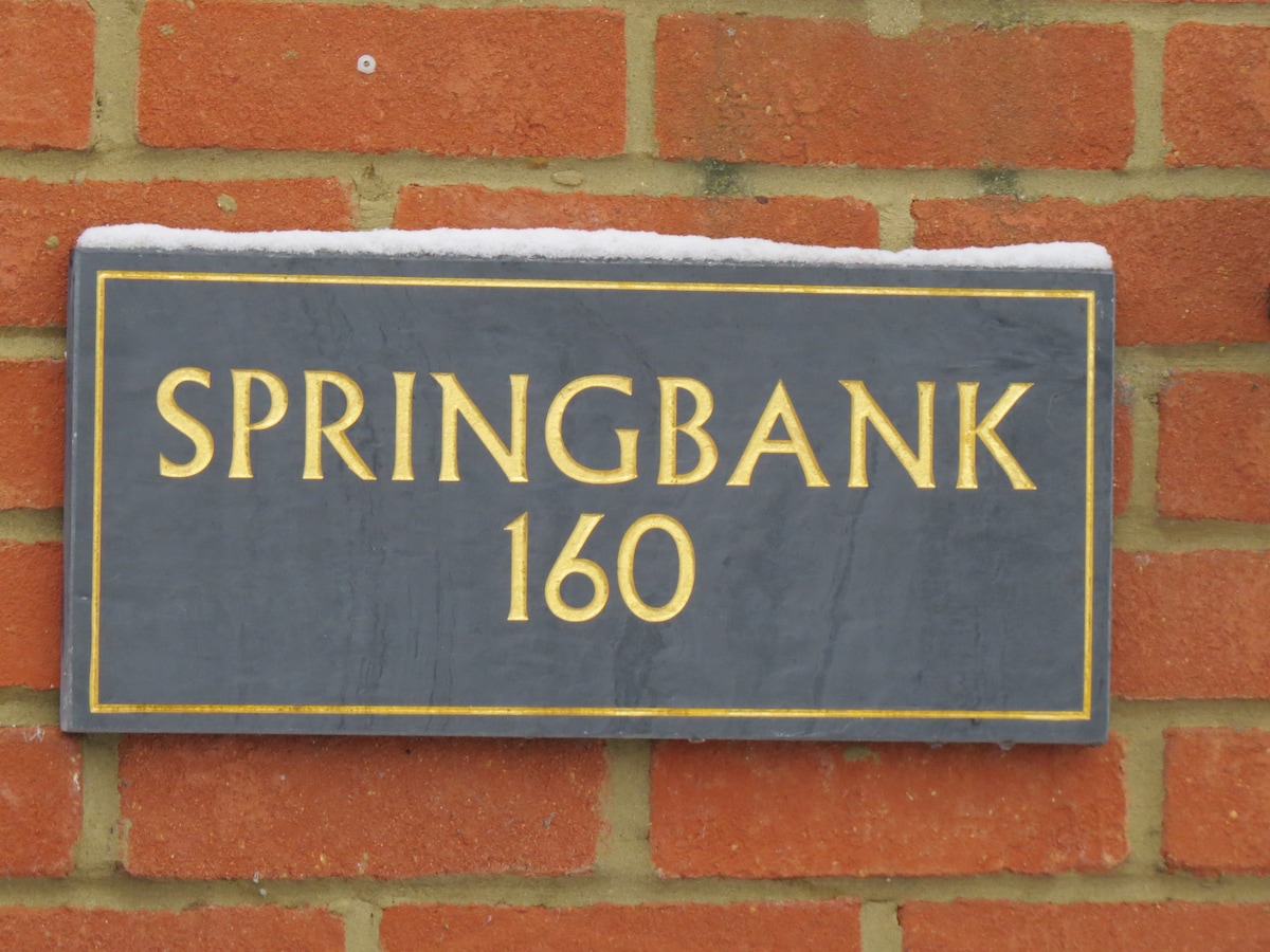 A slate house sign mounted on a brick wall is visible. The sign displays the name 'SPRINGBANK' and the number '160' in golden lettering, contrasting with the dark background. A light dusting of snow sits along the top edge.
