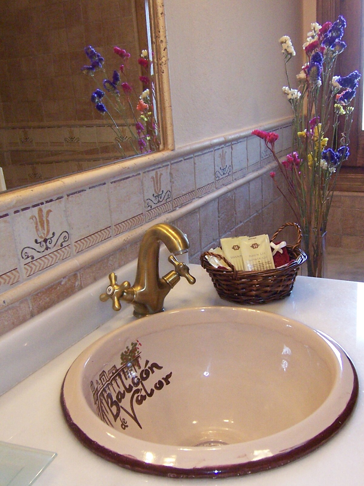 A bathroom sink features a handcrafted bowl with decorative lettering. A vintage-style faucet is positioned above the sink. A small woven basket holds toiletries, while a vase with colorful artificial flowers adds a touch of color to the warm-toned space.