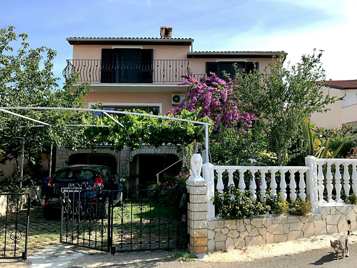 The exterior of a two-story house is surrounded by lush greenery and colorful flowering plants. A secure black gate leads to a driveway with a parked car, and a welcoming white stone fence frames the property. A spacious terrace is visible on the upper level.