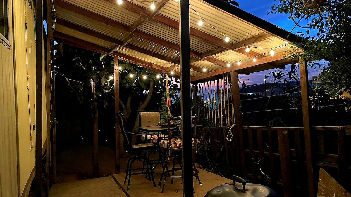 An outdoor patio is featured with a covered area and string lights creating a relaxed ambiance. A small table and chairs are arranged in the center, while the surrounding greenery adds a natural touch. Evening light casts a warm glow over the space.