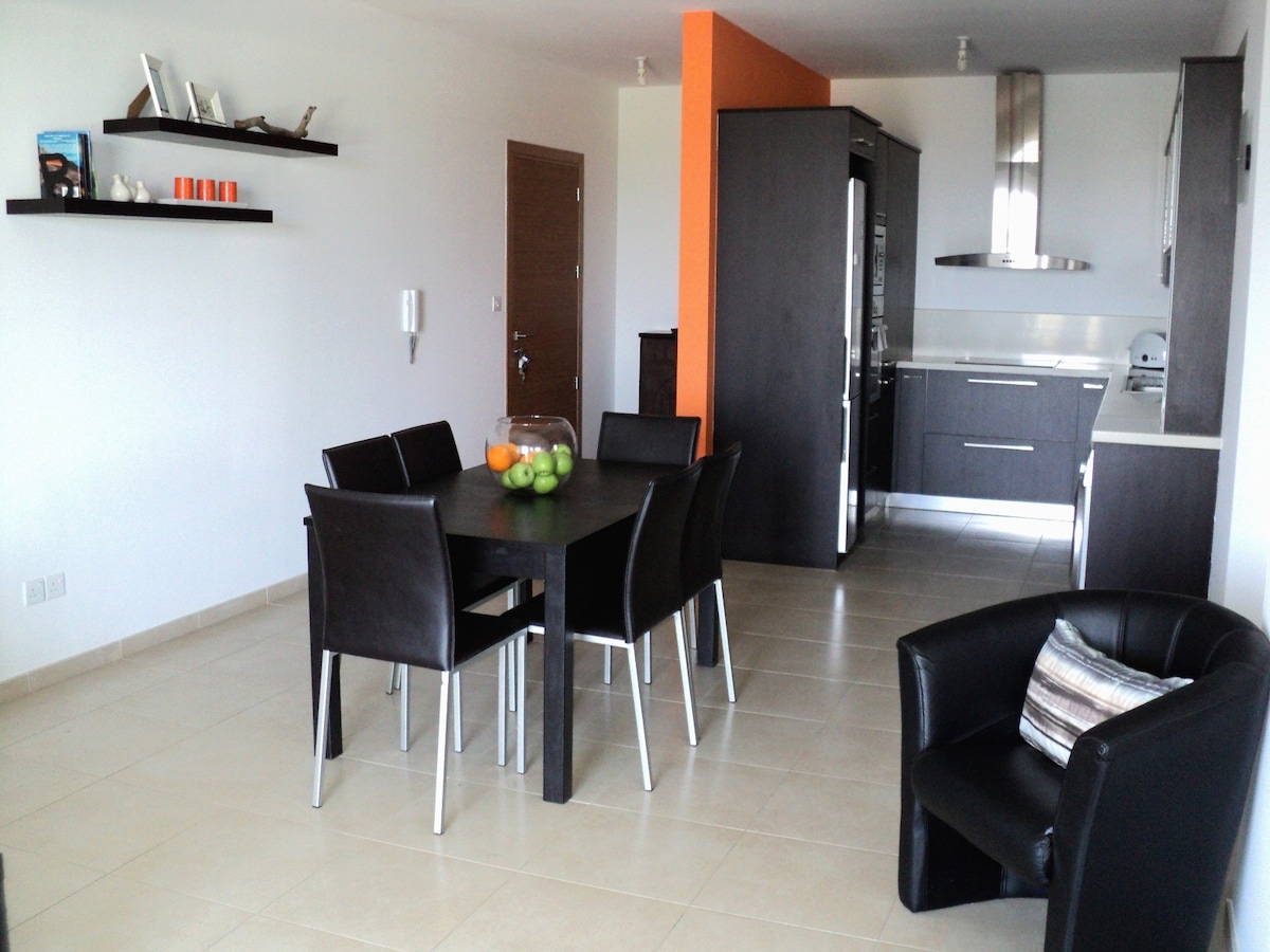 An open-plan living area features a dark wooden dining table surrounded by six black chairs, complemented by a sleek kitchen with modern appliances. A black armchair is positioned nearby, adding to the comfortable ambiance. Decorative shelves with items are mounted on the white walls.
