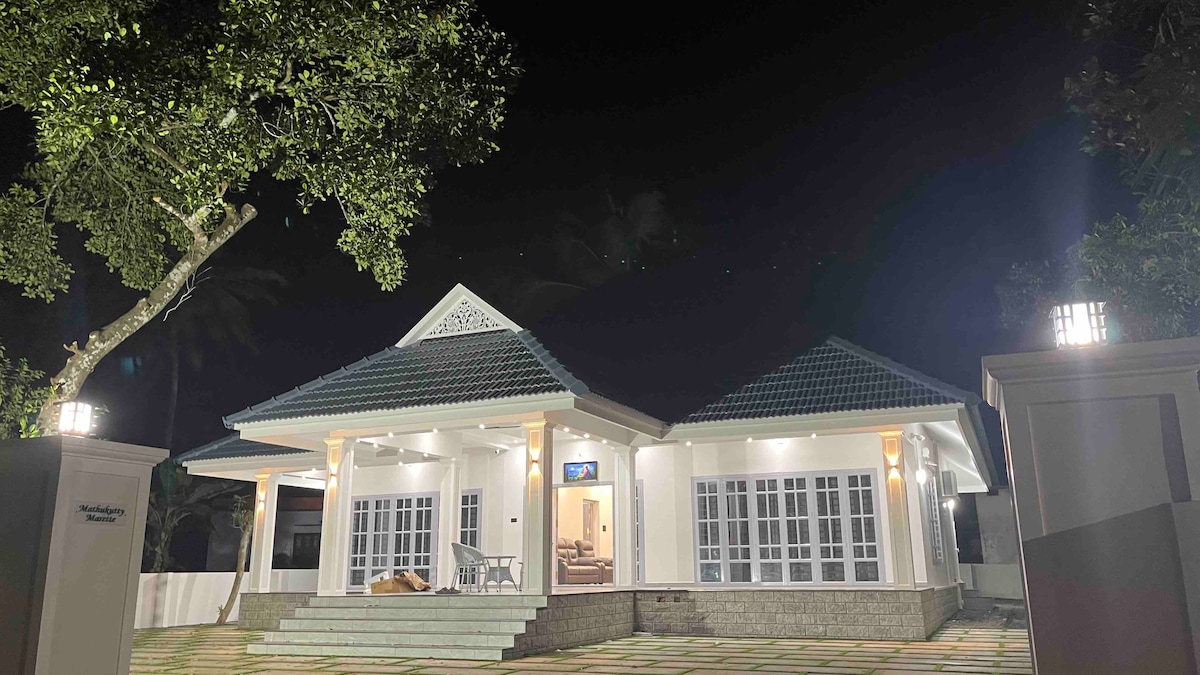 The exterior of the house is illuminated at night, showcasing a modern architectural design. A well-lit porch features multiple entry points with large glass windows, allowing for ample natural light. Lush trees are visible, creating a serene environment.