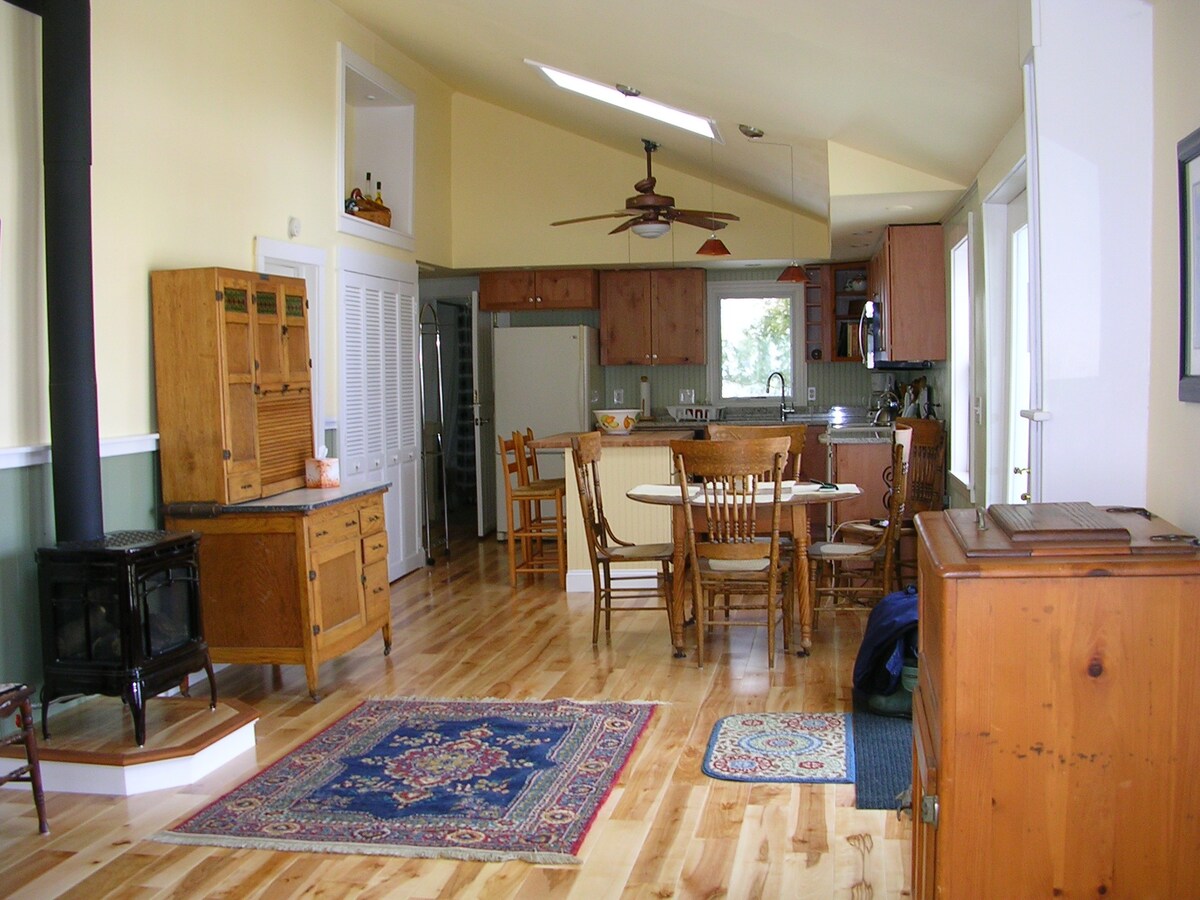 A spacious living area features warm wooden flooring, complemented by a variety of wooden furniture including a dining table with chairs. A rustic stove adds warmth to the room, while natural light enters through multiple windows, enhancing the open layout that leads to the kitchen.
