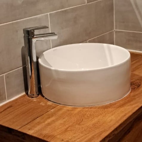 A round white sink is positioned on a wooden countertop. The sleek faucet made of shiny metal complements the modern design. Grey tiled walls provide a subtle backdrop, enhancing the clean, minimalist aesthetic of the bathroom space.