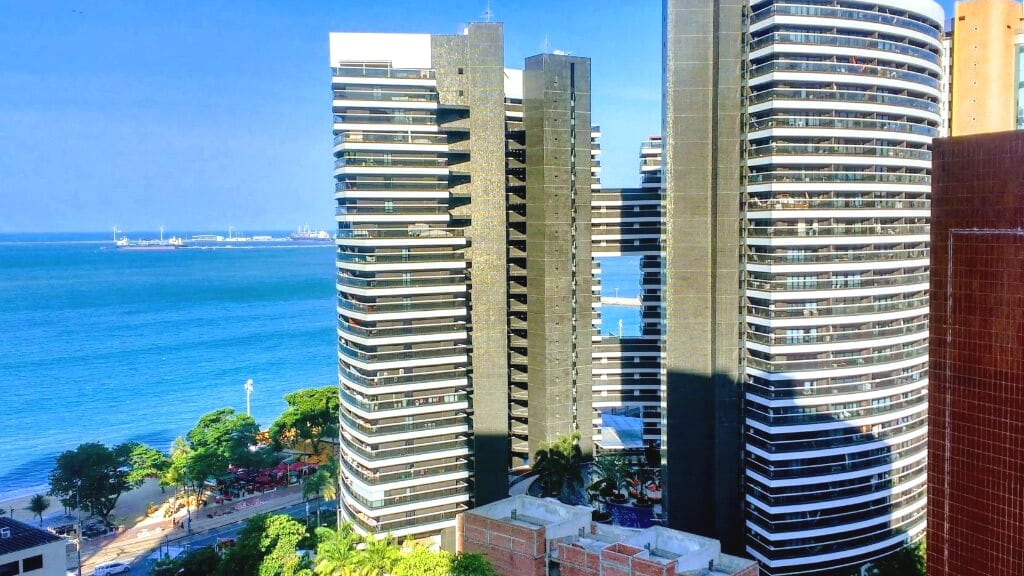Two modern towers with extensive balconies overlook the tranquil blue ocean and sandy beach. Lush greenery surrounds the base of the buildings, while a vibrant beachside promenade is visible in the distance, enhancing the coastal landscape.