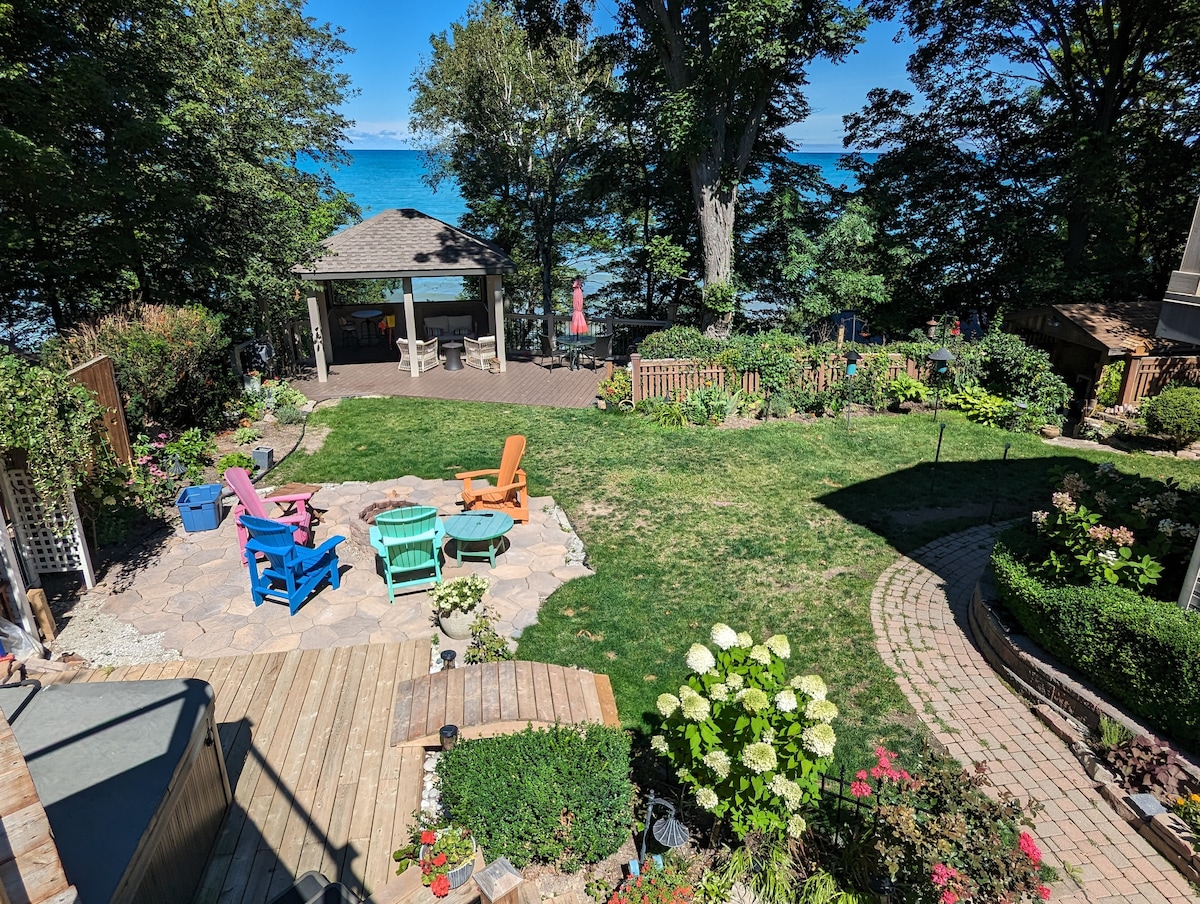 A well-maintained garden area features colorful outdoor seating arrangements, including lounge chairs and Adirondack chairs, surrounded by lush greenery and blooming flowers. A gazebo is visible in the background, overlooking Lake Huron, providing a serene space for relaxation.