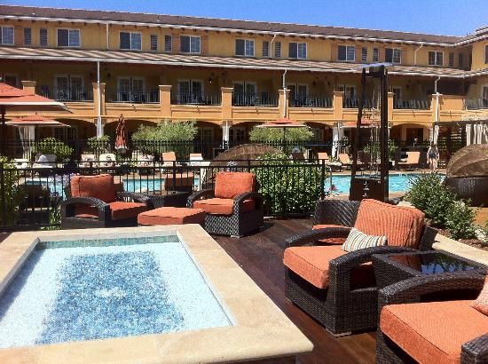 A pool area is surrounded by lounge chairs and shaded umbrellas, inviting relaxation. A hot tub is situated prominently, with plush seating nearby. Lush greenery adds to the space, while the building features multiple balconies overlooking the pool, all under a clear blue sky.