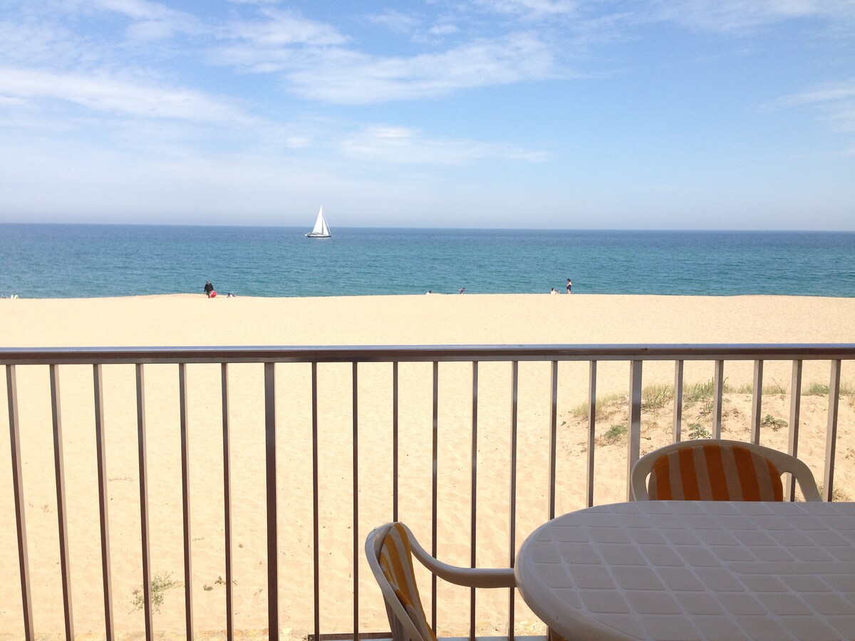 A view from the balcony reveals a sandy beach leading to the calm sea, with a sailboat visible on the horizon. The scene is framed by a railing and a round table with striped chairs, creating a tranquil setting for relaxation.