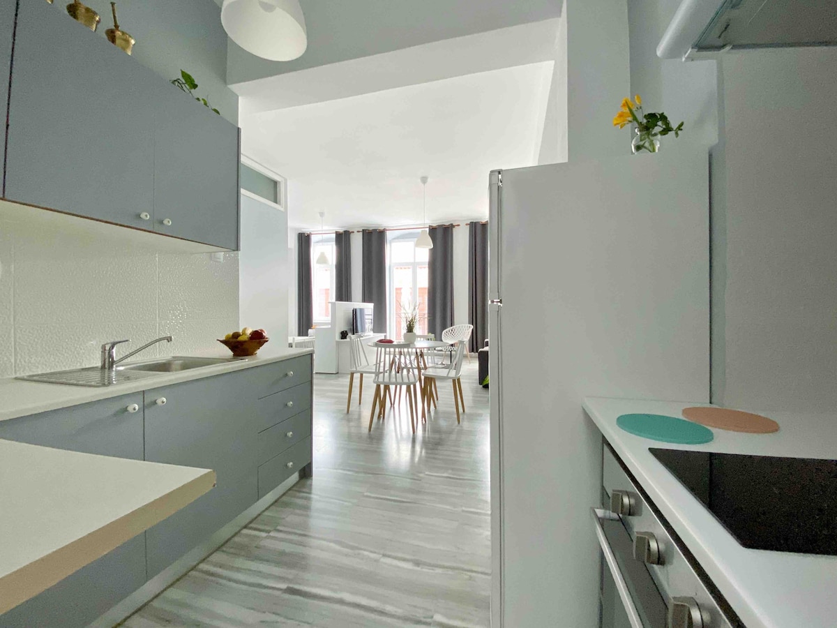 A modern kitchen area is presented with sleek cabinetry and a neutral color palette. The space features a sink and stove, while a bright dining area is visible in the background, showcasing a table surrounded by stylish chairs and large windows allowing ample natural light.