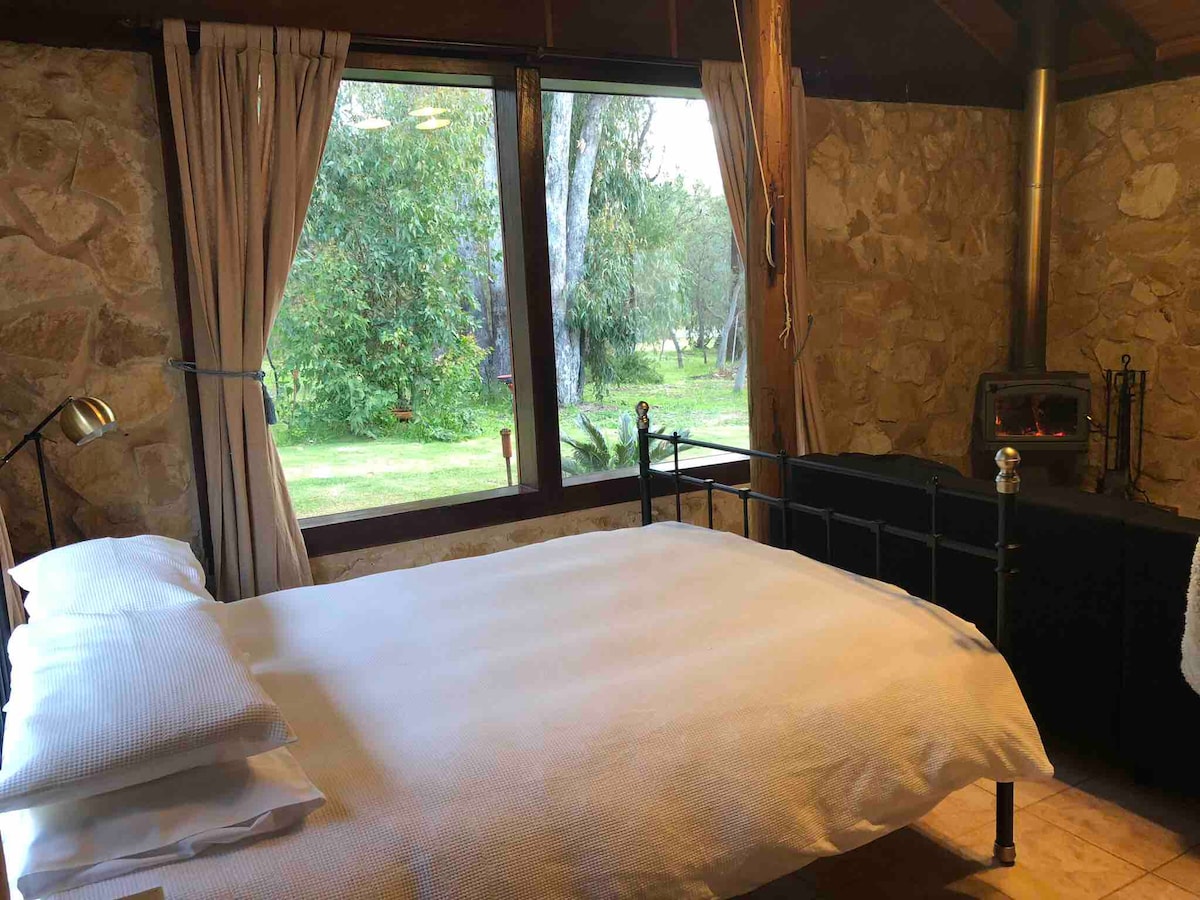 The interior of the cottage features a spacious bed with crisp white linens, positioned adjacent to a large window offering views of the natural bushland. A cozy pot-belly stove is visible, surrounded by stone walls and soft curtains that frame the outdoor scenery.