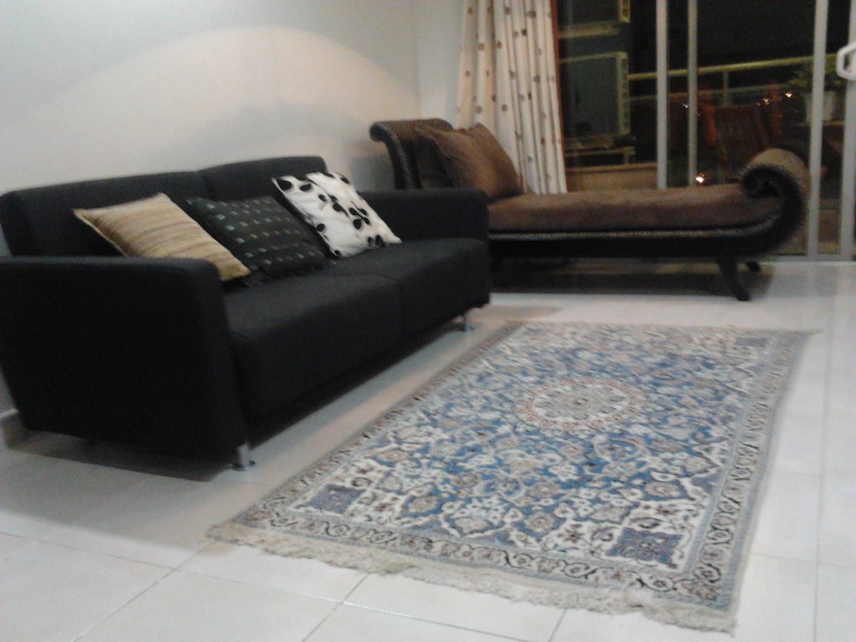 A comfortable living room features a dark sofa adorned with decorative pillows and a chaise lounge. A patterned area rug covers the tiled floor, adding a touch of warmth. Large glass doors provide ample natural light and access to the balcony.