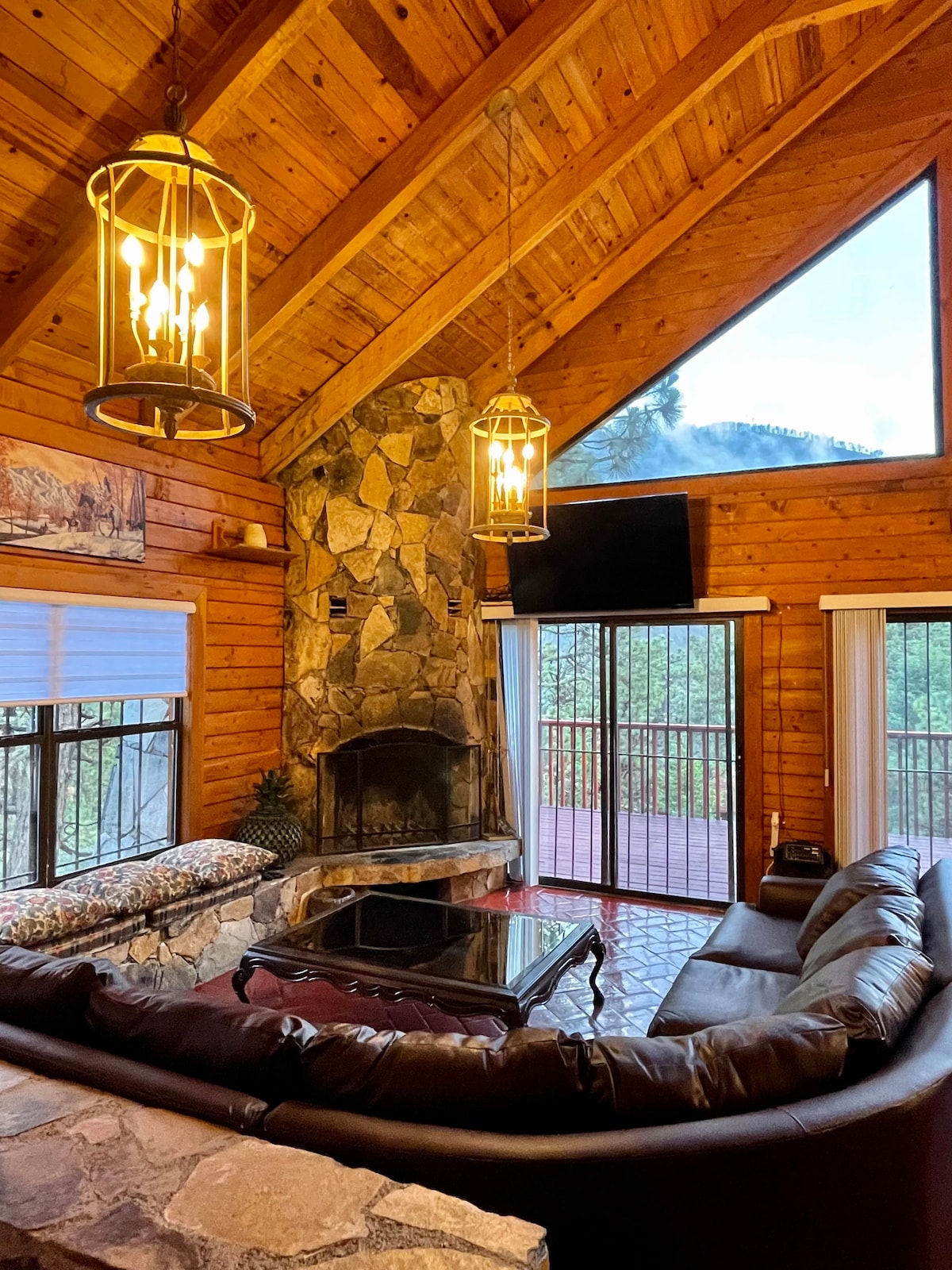 The living room features a stone fireplace as the focal point, complemented by wooden beams and expansive windows that allow natural light. A leather sectional sofa wraps around a coffee table, while pendant lights hang from the high ceiling, creating a cozy gathering space.