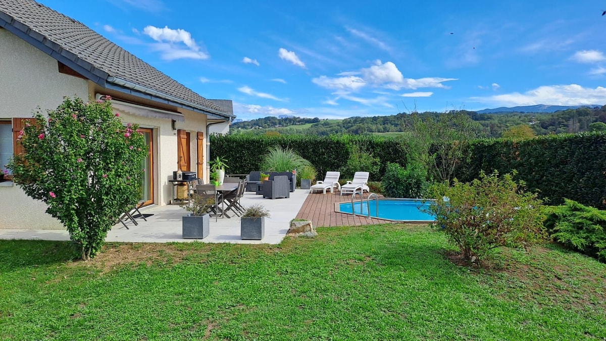 A serene outdoor area is shown, featuring a swimming pool surrounded by lounge chairs. A dining set is positioned on a wooden deck, while vibrant greenery and a hedge form a natural boundary. The house is adorned with large windows, allowing natural light to fill the space.