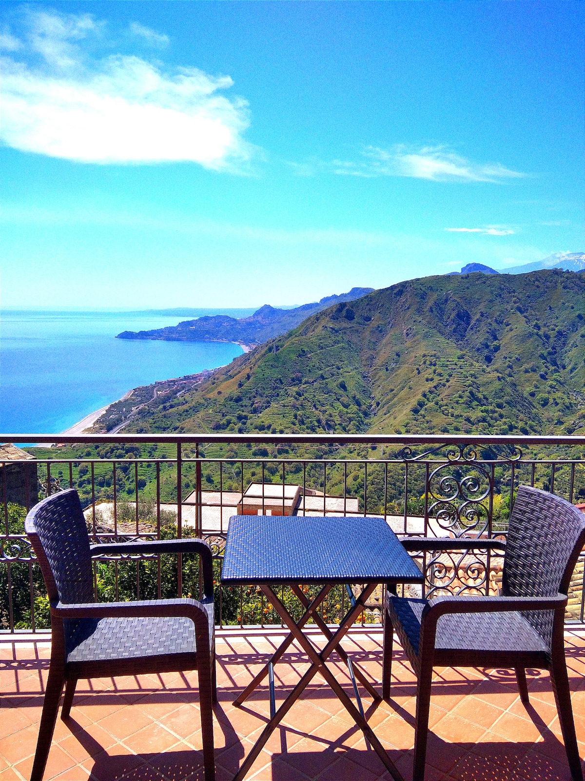 A small outdoor terrace features a round table and two chairs, positioned against a stunning backdrop of hills and a distant coastline. The expansive blue sky compliments the lush greenery, creating a serene setting for relaxation.