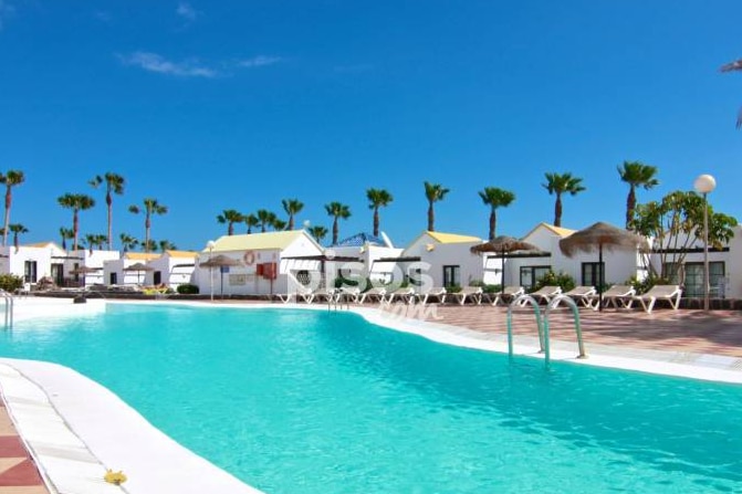 A large swimming pool is surrounded by lounge chairs and palm trees, creating a relaxing outdoor area. The pool's clear blue water reflects the bright sky, and white buildings with yellow roofs are visible in the background.