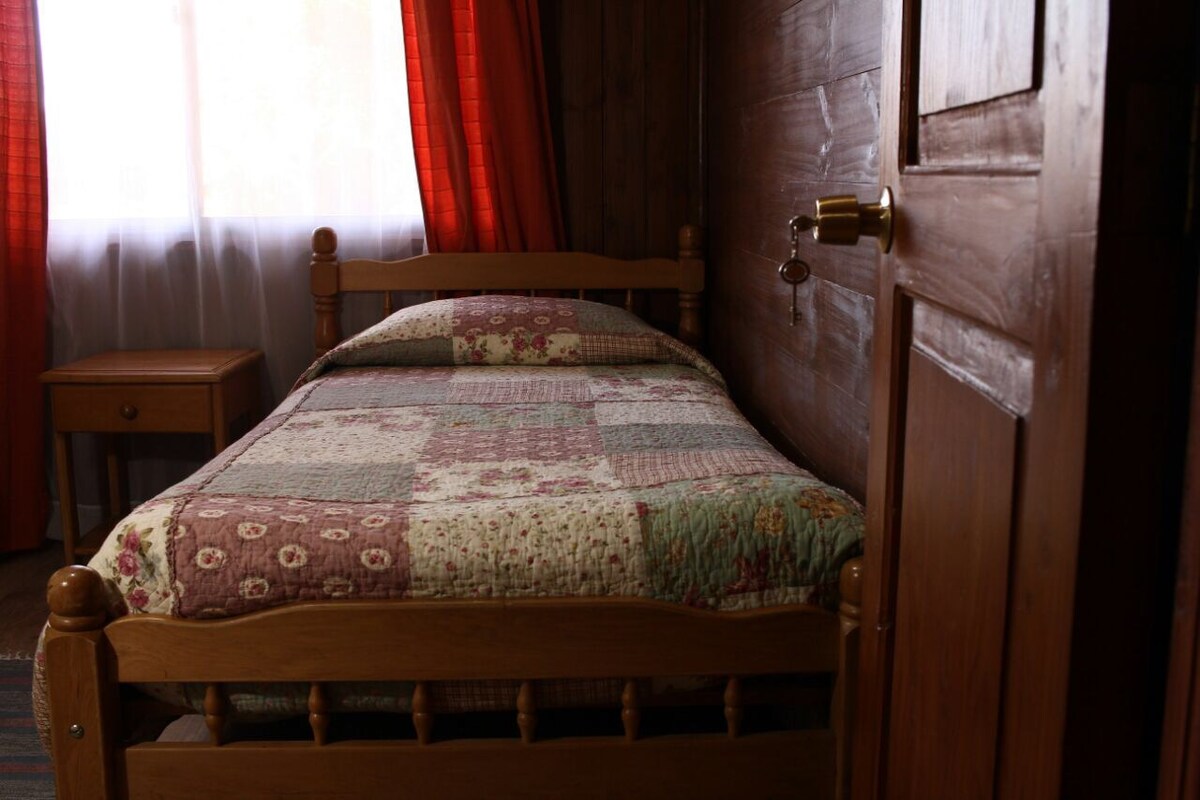 A cozy sleeping area is furnished with a single bed dressed in a patchwork quilt. Light filters softly through a window dressed with sheer curtains, complemented by red drapes. A wooden side table is situated beside the bed, enhancing the warm wooden interior.