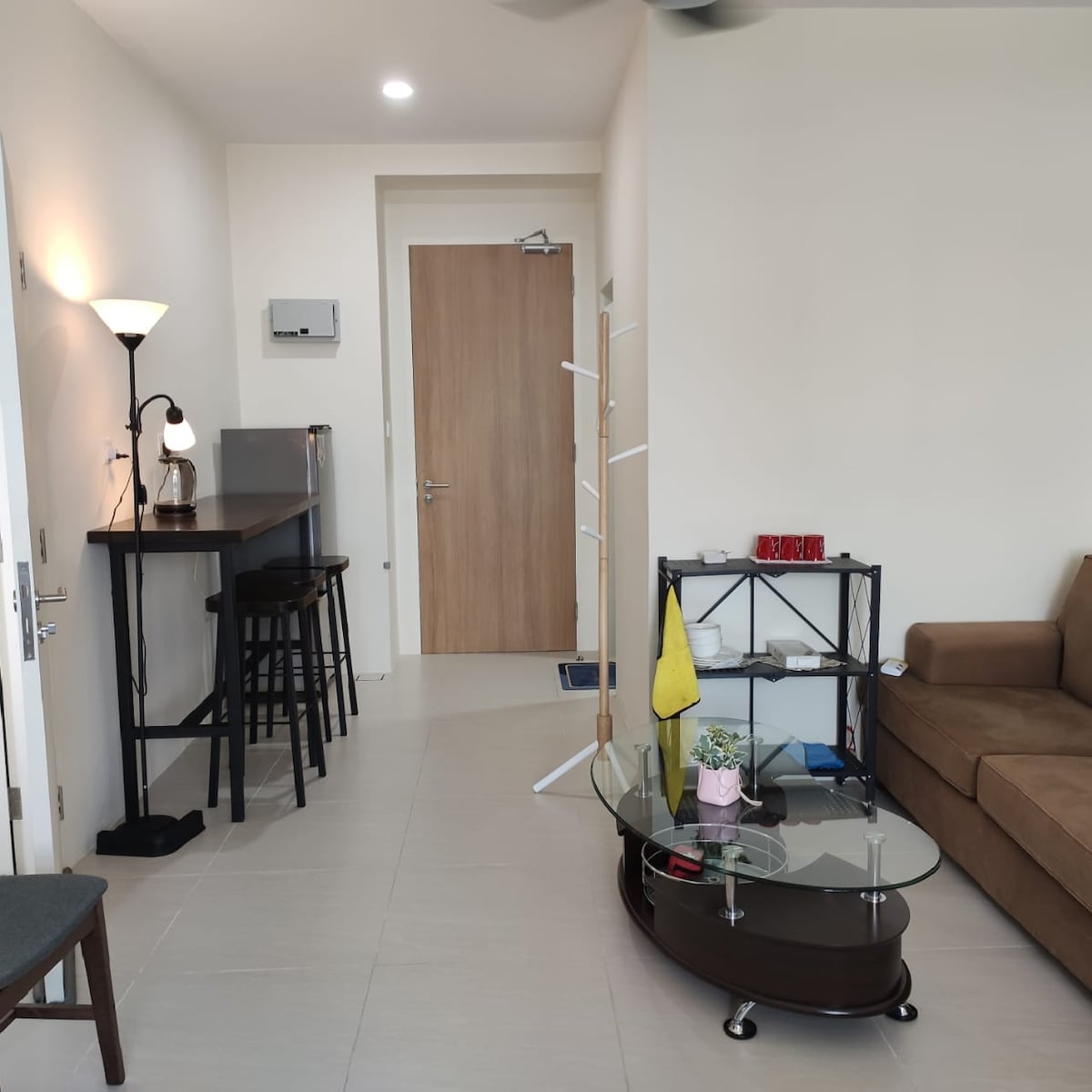 A modern living area features a comfortable brown sofa paired with a glass coffee table. A bar-style dining table with high chairs is positioned adjacent to a kitchenette. Bright lighting is provided by a standing lamp, while a door leads to the entrance of the condo.