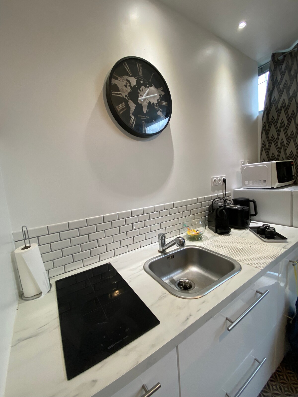 The kitchen area features a modern layout with a marble countertop, equipped with a sink and vitro ceramic cooktop. A microwave, kettle, and coffee maker are also visible, along with a wall clock. Subway tile backsplashes add a sleek touch to the space.