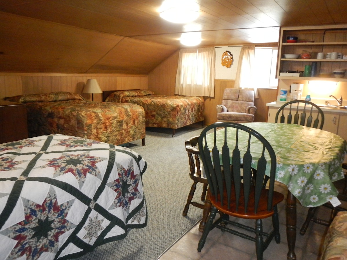 The cabin's interior features three full-size beds and one twin bed adorned with patterned bedding. A kitchenette is visible alongside a small dining table with wooden chairs. Natural light enters through the window, illuminating the warm wood paneling and cozy atmosphere.