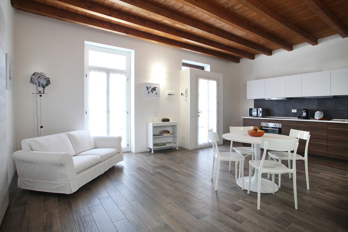 The open living space is designed with wooden ceiling beams, featuring a white sofa and a round dining table with white chairs. Large windows allow natural light to fill the area, complementing the modern kitchen with wooden cabinetry and essential appliances.