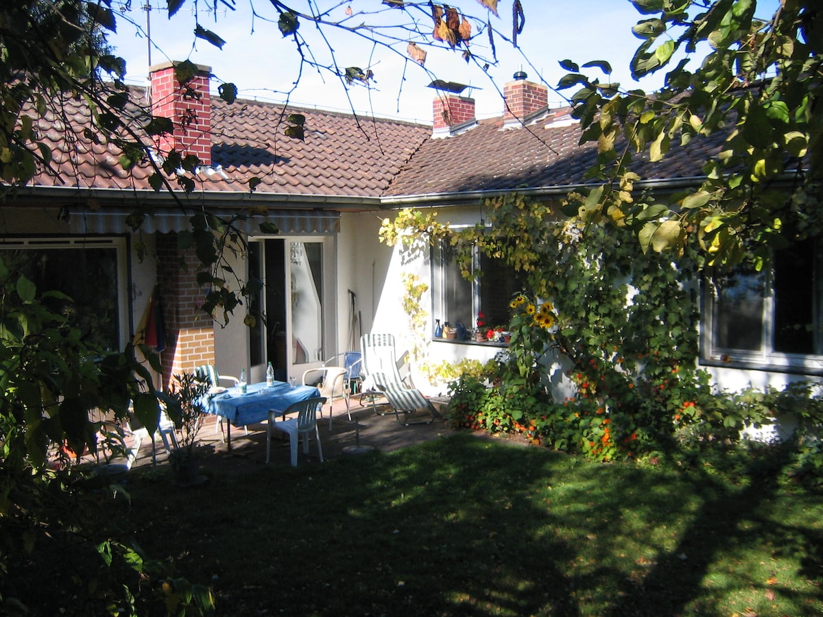 A charming bungalow exterior is seen under a clear blue sky, with a patio area featuring a table and chairs. Lush greenery surrounds the space, including vibrant flowerbeds and sunflowers. Large windows provide views into the interior, enhancing the connection to the outdoors.
