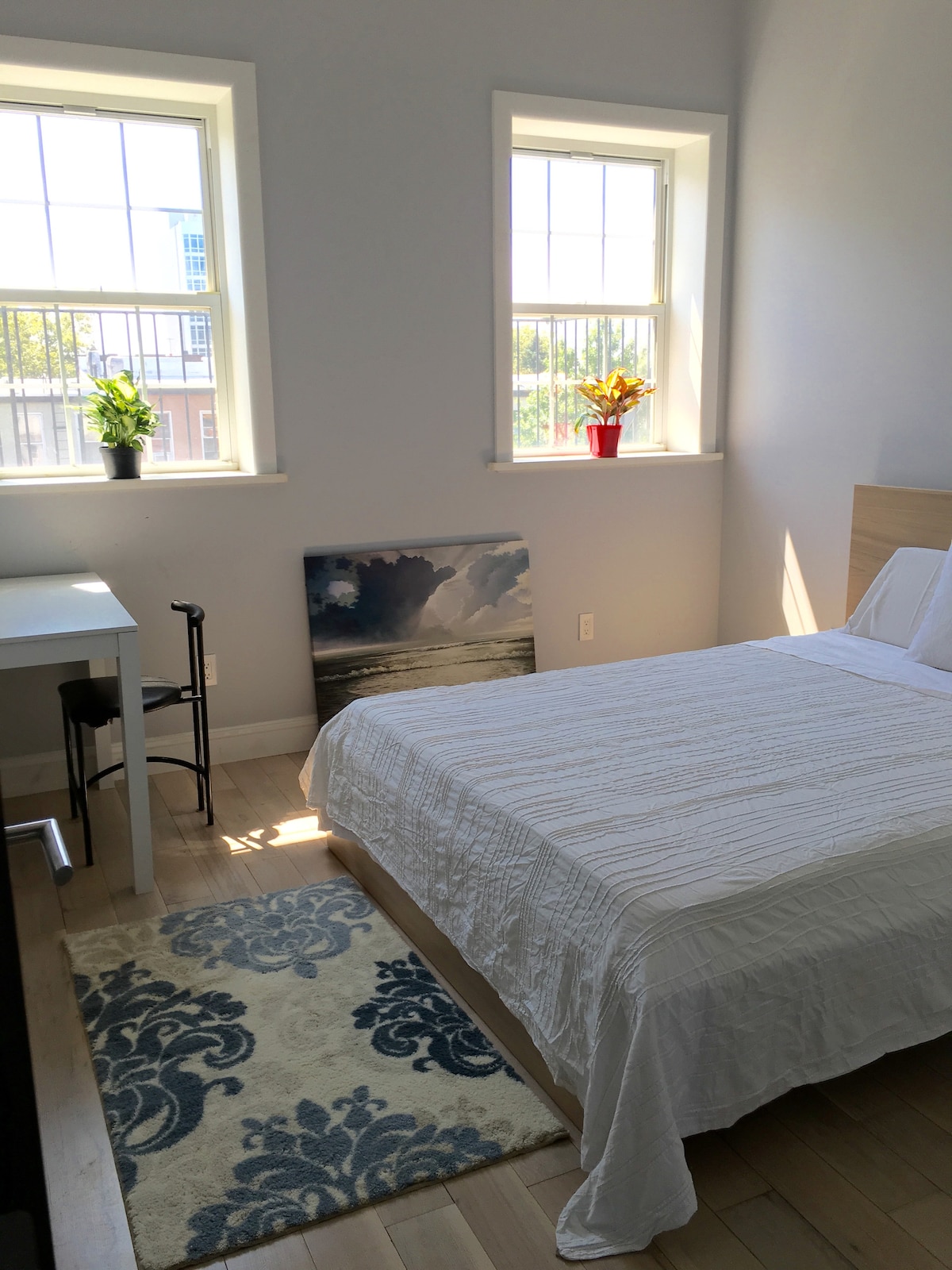 A bright, airy bedroom includes two windows allowing natural light to illuminate the space. A spacious bed is covered with a white quilt, while a small desk and chair are positioned nearby. A decorative area rug and a piece of wall art enhance the room's inviting feel.