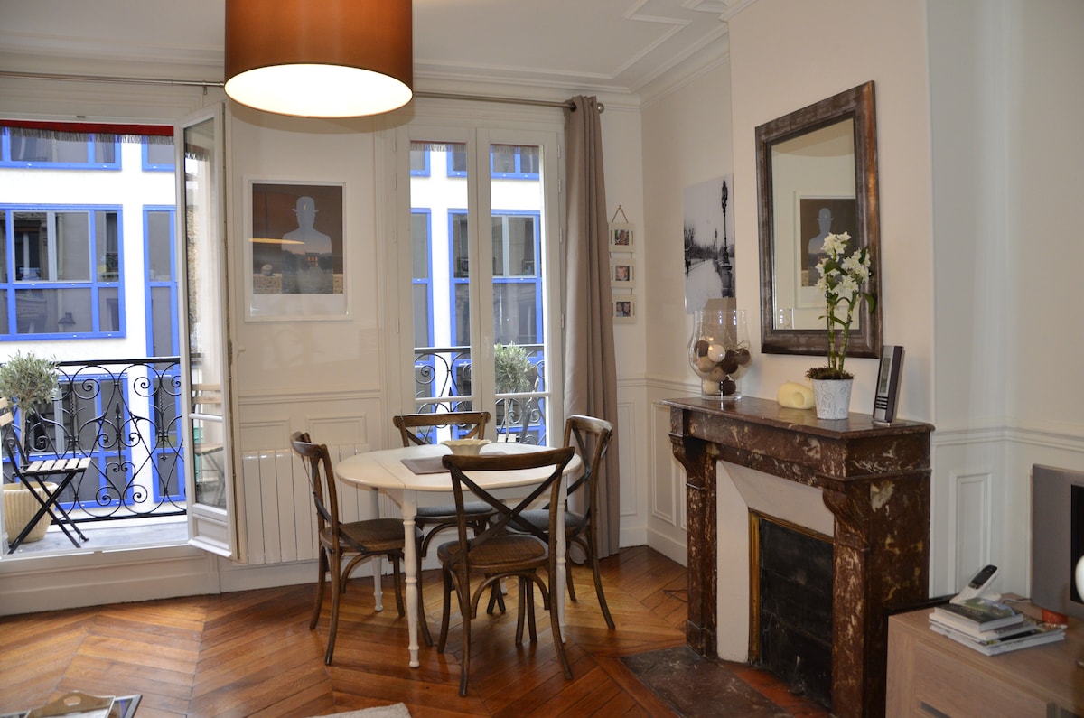 A light-filled dining area features a round table surrounded by wooden chairs. Large French doors open onto a balcony, allowing natural light to fill the space. A decorative fireplace adds character, complemented by a mirror and a small vase with flowers on display.