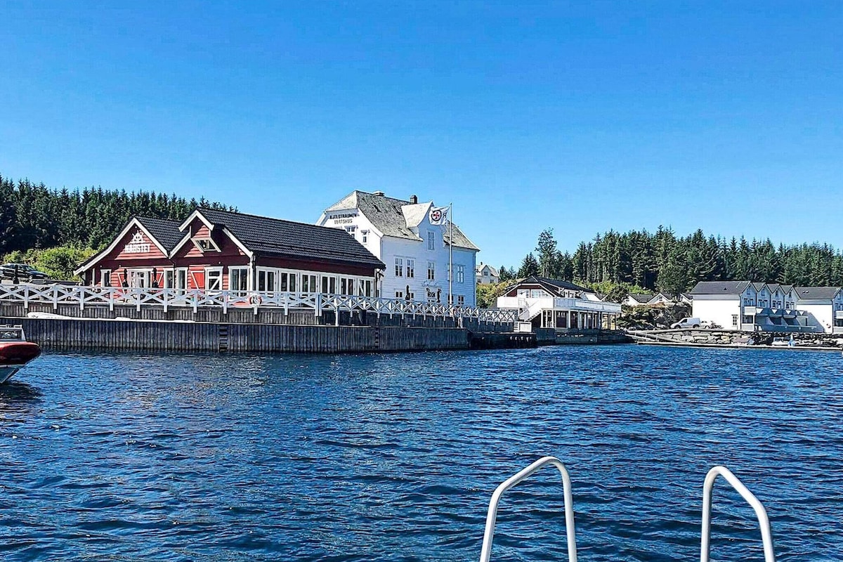 A waterfront view of a colorful fisherman's cabin alongside other buildings, created with a mix of traditional and modern architecture. The calm blue water reflects the sunlight, and lush greenery is visible in the background, enhancing the coastal scenery.