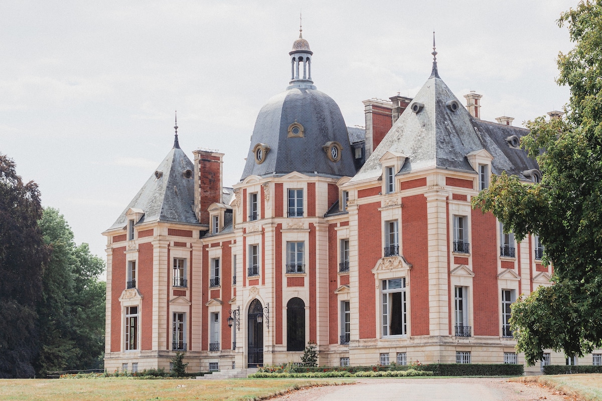 The elegant chateau features a striking architectural design, characterized by its red brick facade and elaborate roofing. Towering domes and intricate details enhance its regal appearance, surrounded by manicured grounds that provide a picturesque setting.