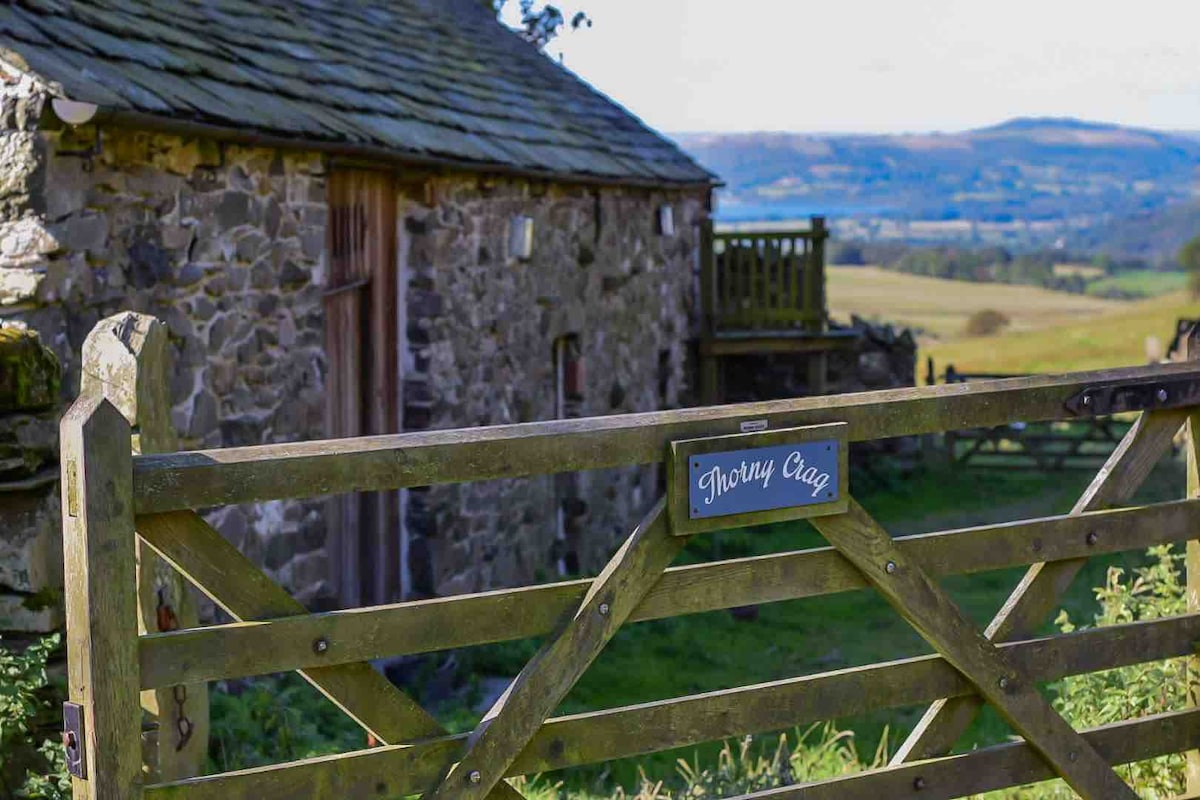 A rustic wooden gate is featured, marked with a sign that reads 'Thorny Crag.' The background showcases a stone barn with large windows, set against a hillside with expansive views of the landscape beyond.